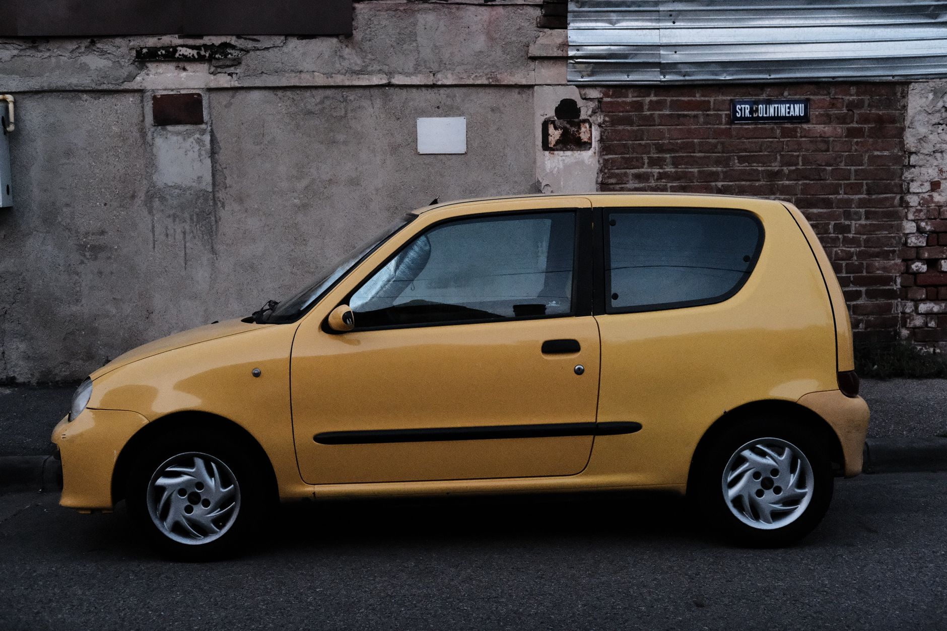 Side view of a parked yellow Fiat Seicento on a city street with a rustic wall backdrop.