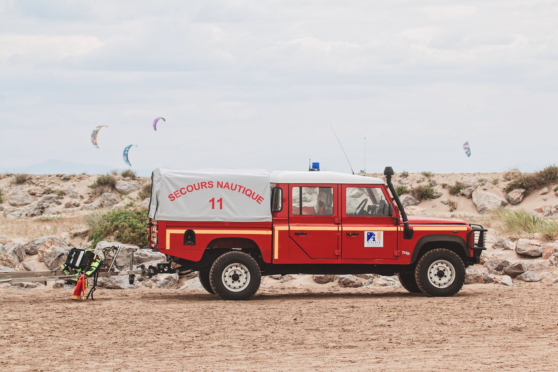 Red rescue truck parked on sandy beach with kite surfers in background, Secours Nautique visible.