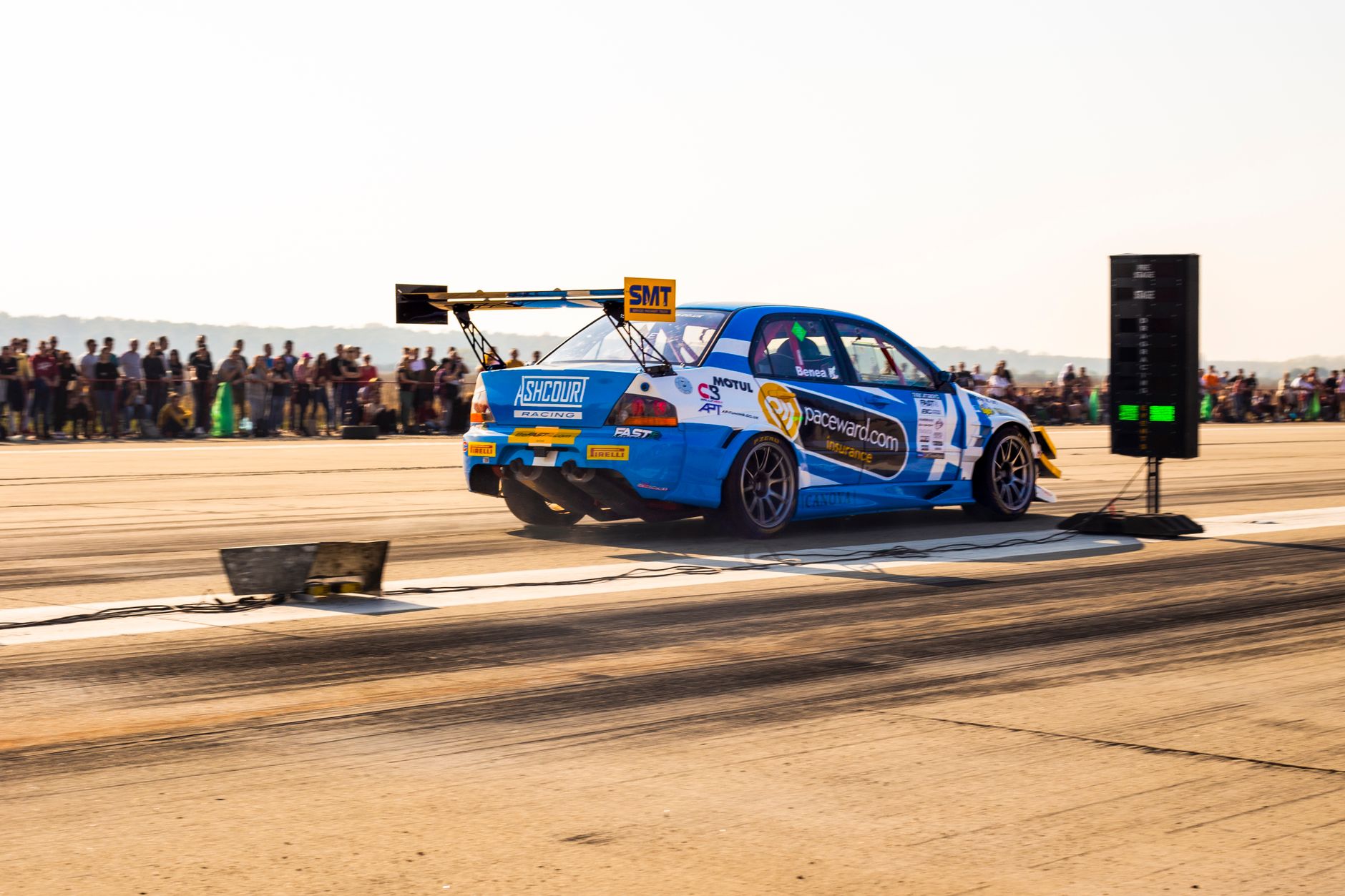 Exciting drag racing scene with a blue race car speeding past the starting line in front of a crowd.