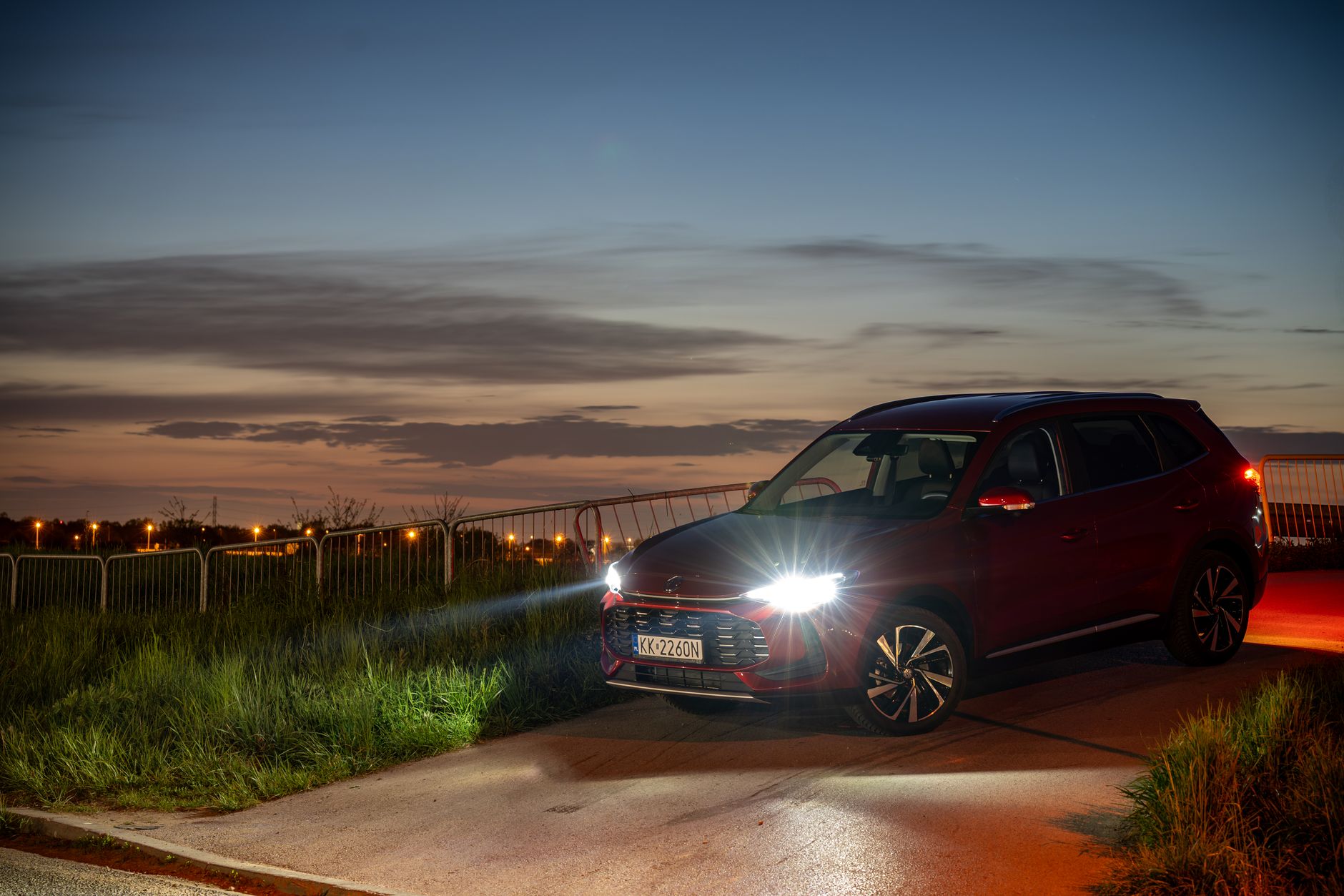 Red SUV with headlights on parked on a country road during twilight in Kokotów, Poland.
