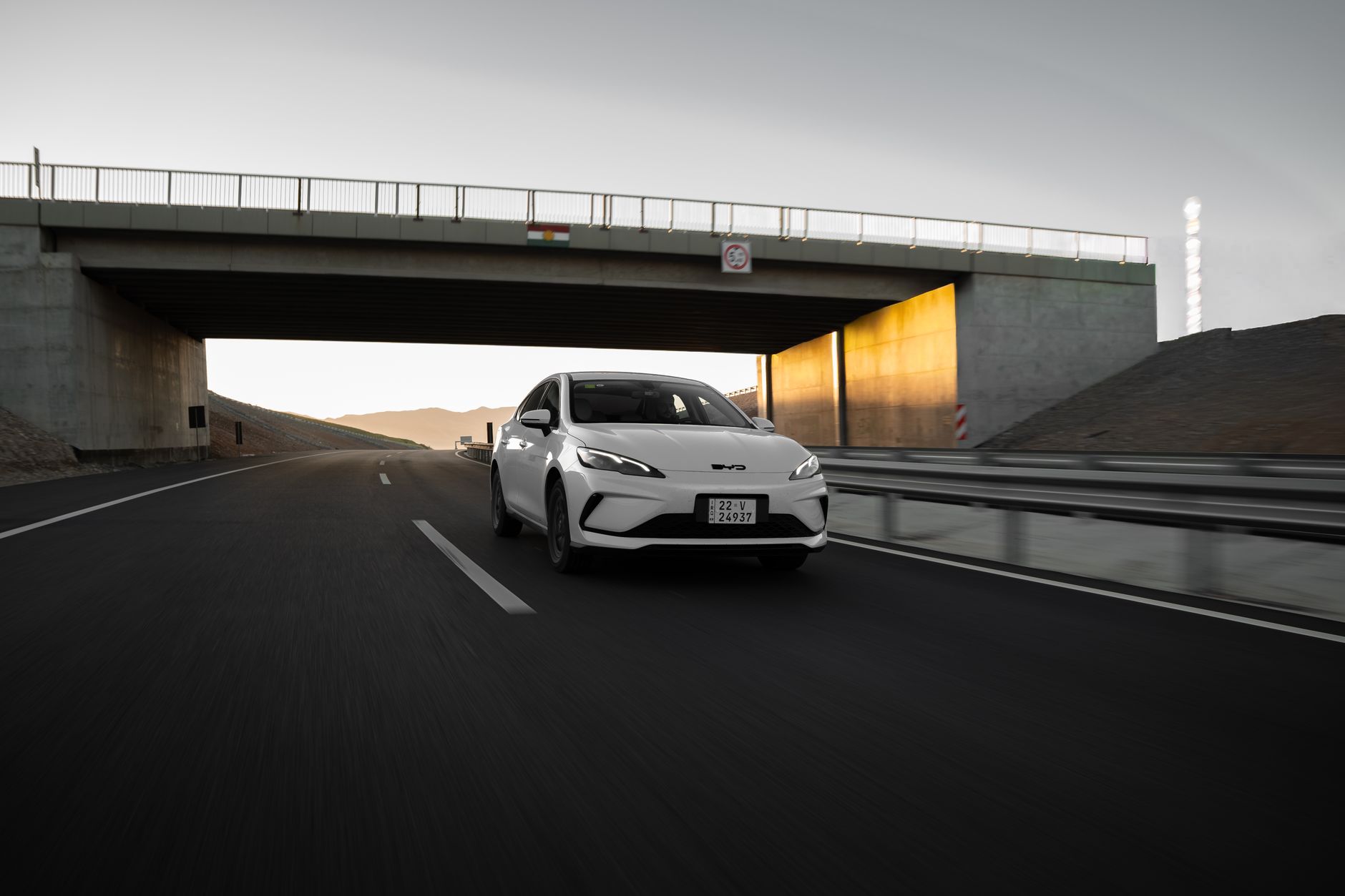 A white modern car driving under a bridge on a highway at sunset, showcasing speed and elegance.