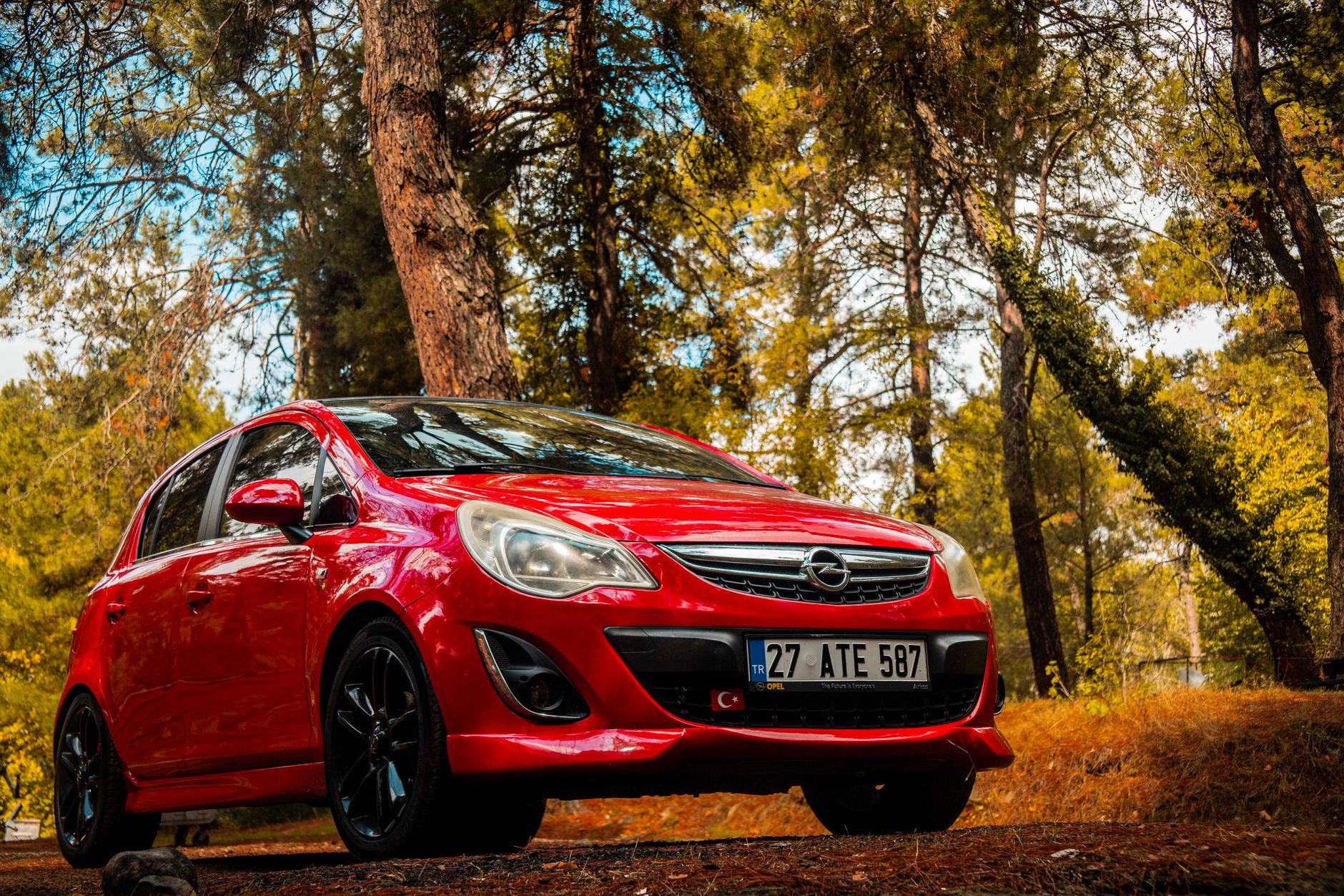 A vibrant red Opel Corsa parked amidst a serene autumn forest located in Kozak, İzmir, Türkiye.