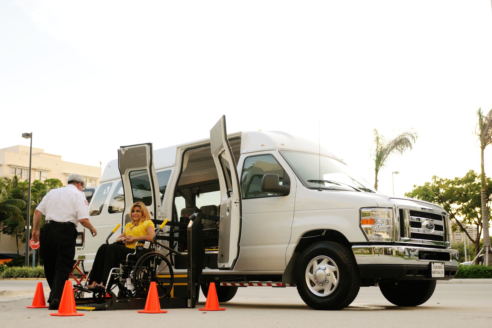 A person in a wheelchair uses a lift to board a van, assisted by a driver. Outdoor setting with cones.