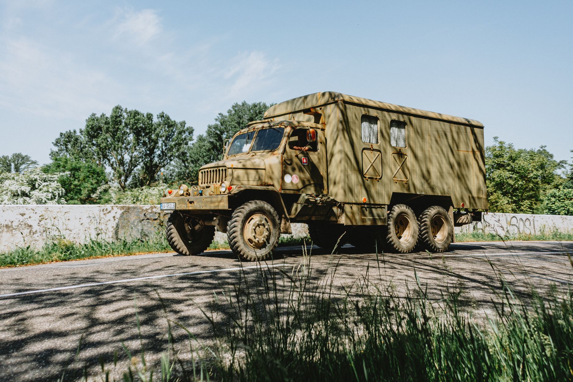 An old military truck driving on a rural road surrounded by greenery in summer.