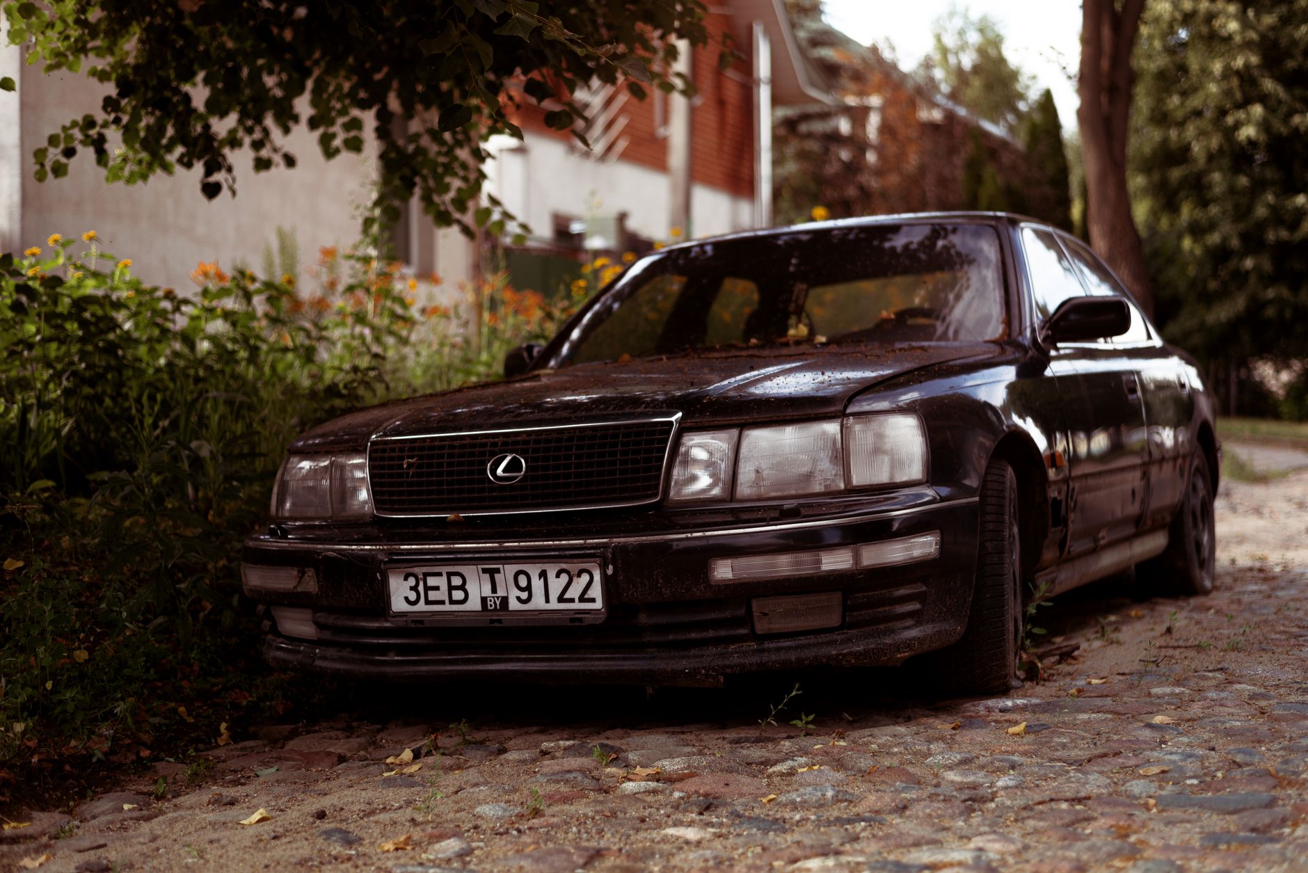 A classic Lexus LS in black livery parked on a cobblestone street surrounded by lush greenery.