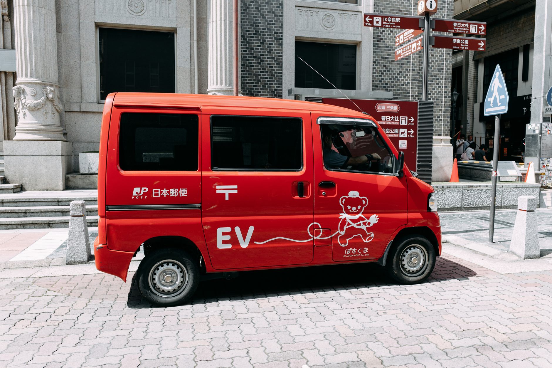 A red electric delivery van parked on a street in an urban area with directional signs.