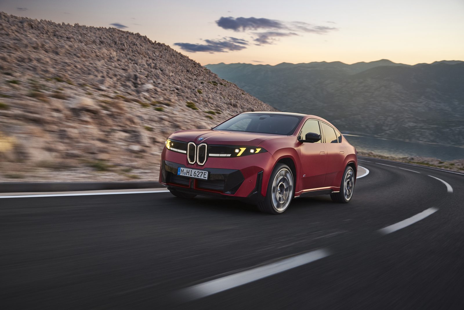 Czerwone BMW iX4 M60 xDrive podczas jazdy krętą górską drogą o zachodzie słońca.