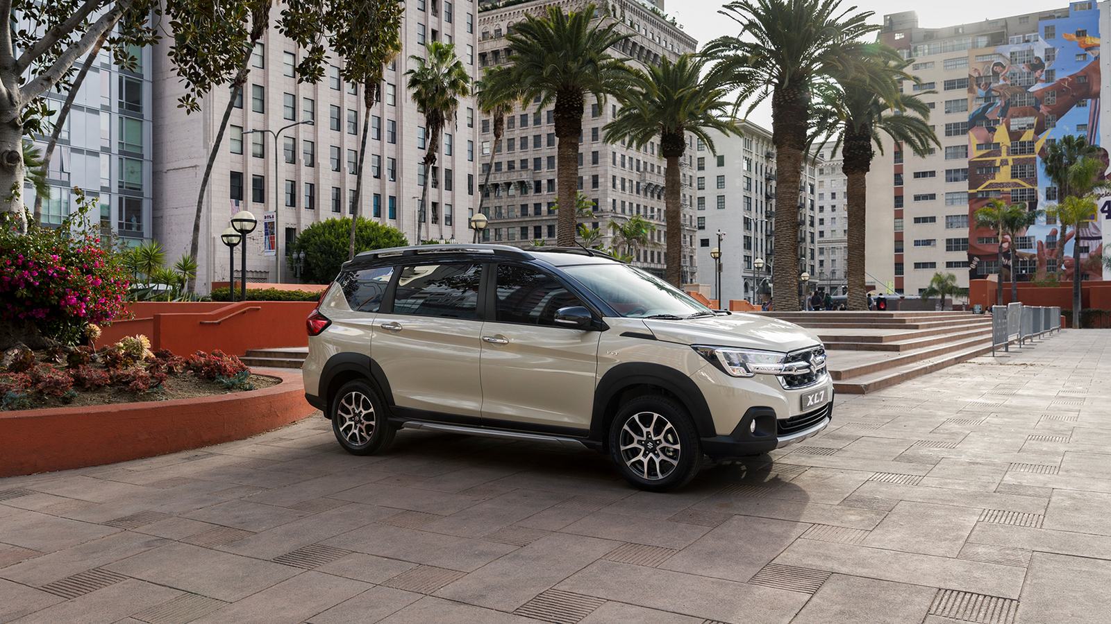 Beżowe Suzuki XL7 zaparkowane na miejskim placu wśród palm i wysokiej zabudowy.