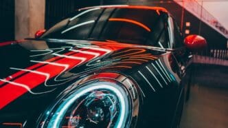 Close-up of a black sports car with vibrant red and white neon light reflections, parked in an urban garage.