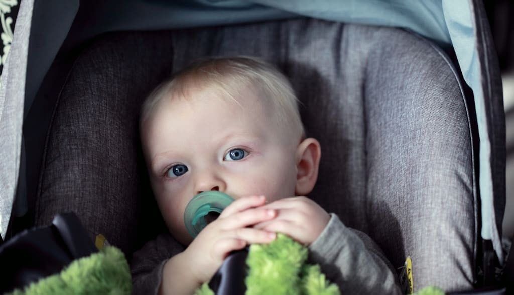 Blond niemowlę z niebieskimi oczami siedzące w foteliku samochodowym z zielonym kocykiem i smoczkiem — pytanie: kiedy kończy się obowiązek jazdy w foteliku samochodowym?