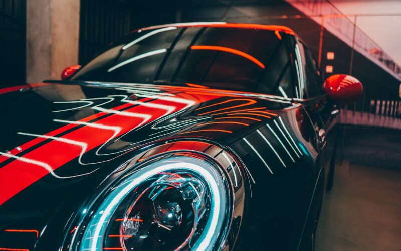Close-up of a black sports car with vibrant red and white neon light reflections, parked in an urban garage.