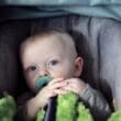 Blond niemowlę z niebieskimi oczami siedzące w foteliku samochodowym z zielonym kocykiem i smoczkiem — pytanie: kiedy kończy się obowiązek jazdy w foteliku samochodowym?