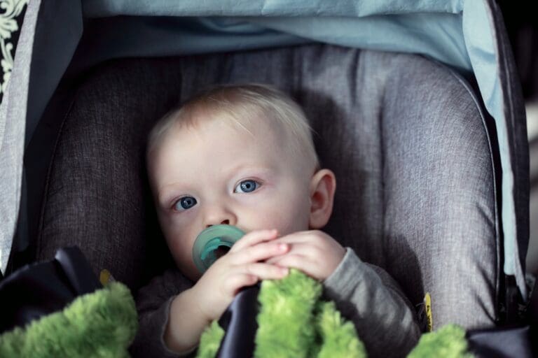 Blond niemowlę z niebieskimi oczami siedzące w foteliku samochodowym z zielonym kocykiem i smoczkiem — pytanie: kiedy kończy się obowiązek jazdy w foteliku samochodowym?